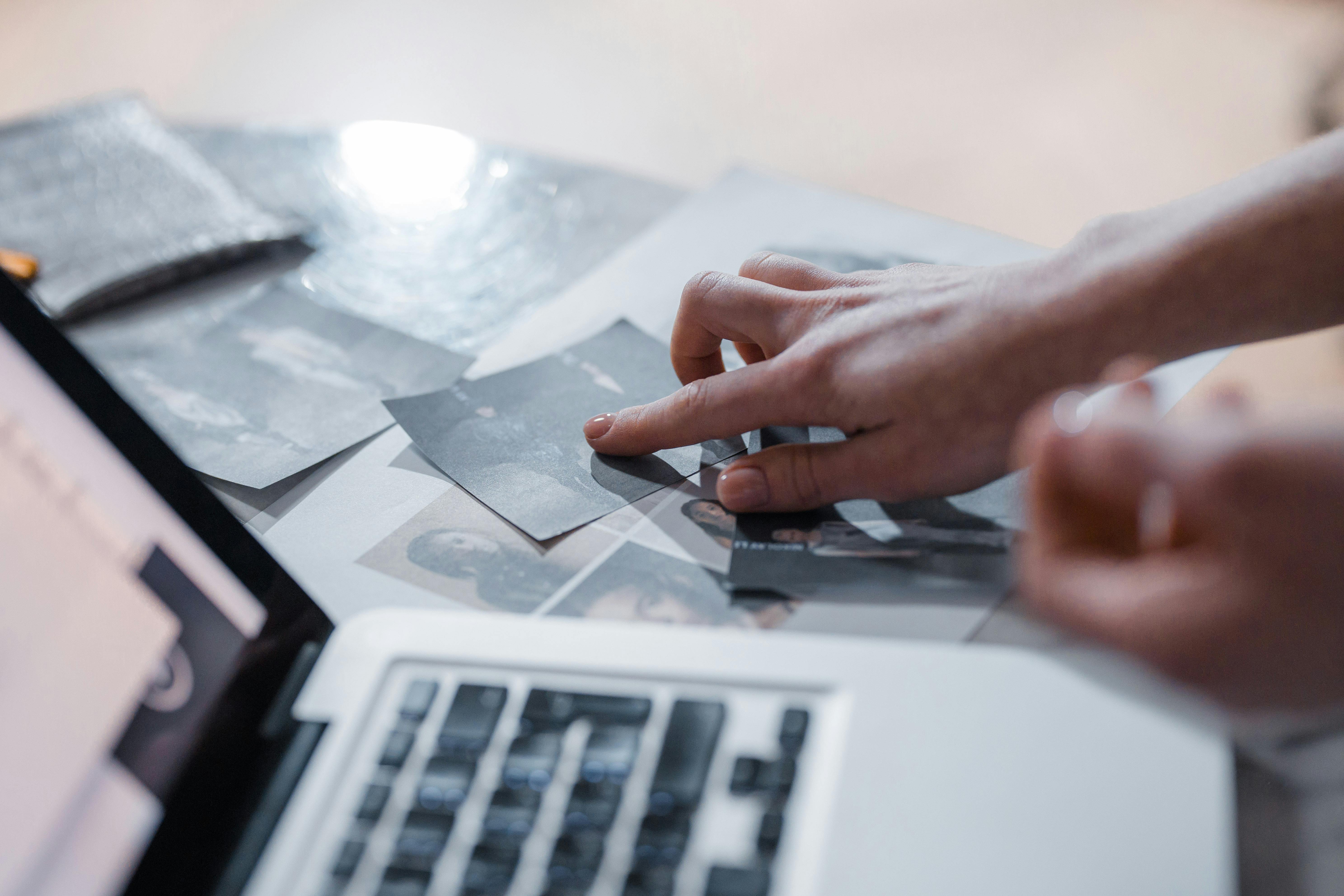 Picture of an open laptop next to old photographs, with a hand pointing at one of the photos