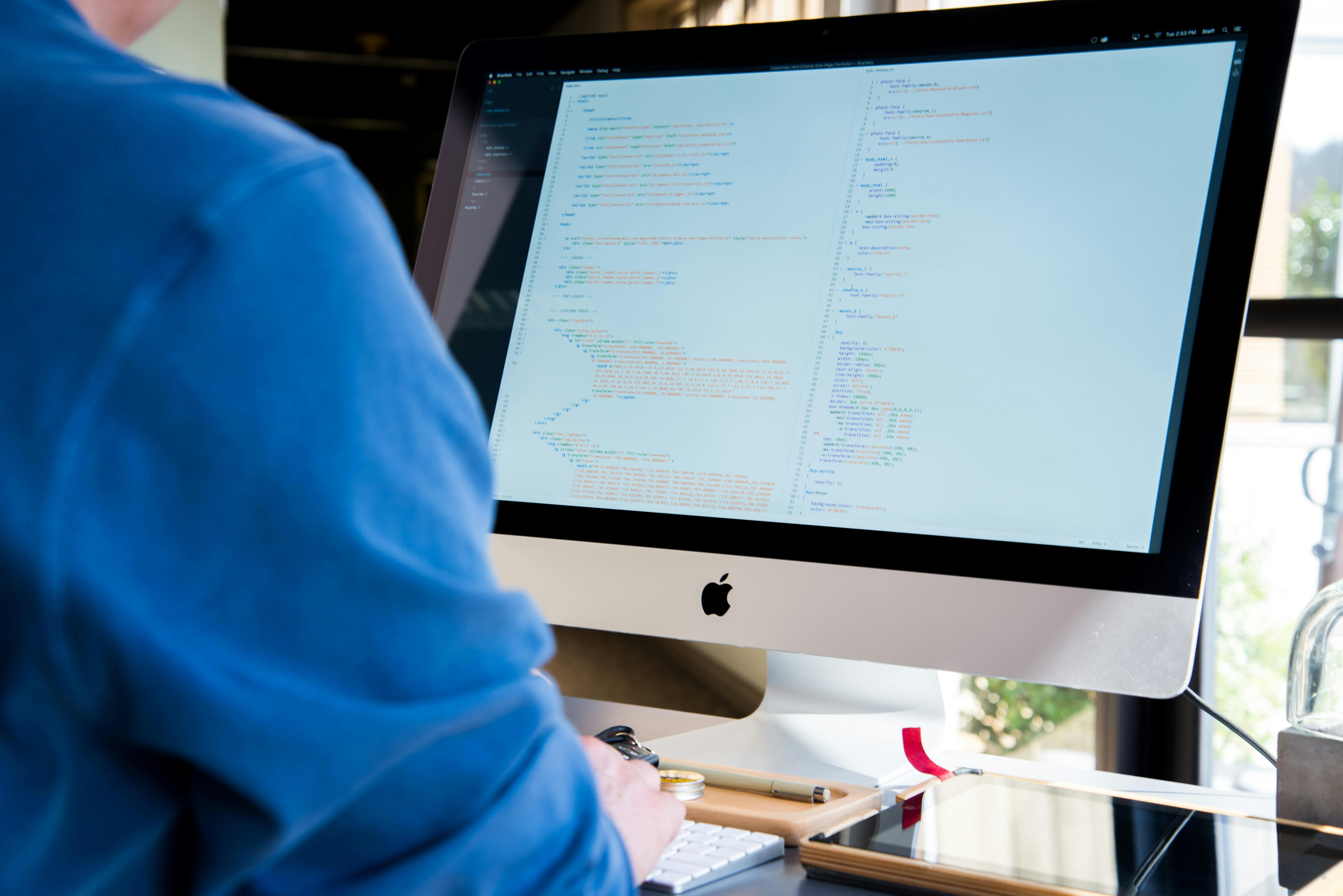 Image of a computer screen with coding displayed and a person wearing a blue shirt facing the computer with their hands on a keyboard